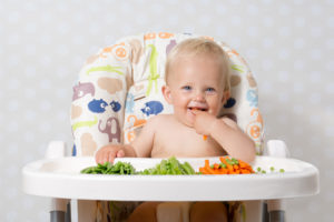 A baby smiles in their feeding chair while reaching for veggies.