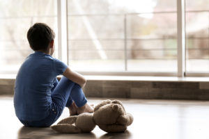 A young boy sits on the floor and stares out the window. He appears upset and sits next to a stuffed animal.