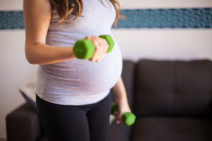 A pregnant woman lifts green dumbbell weights inside her home.