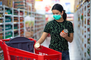 A person wears a medical mask while they grocery shop. They put two canned items into their grocery cart inside the grocery store.