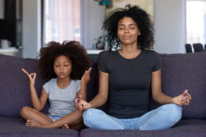 A mother and a daughter meditate in their living room as they sit on a couch.