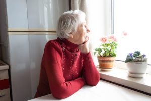 An elderly woman wearing a red sweater sits at a table and stares out a window longingly. She appears to be daydreaming.