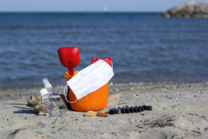 An orange sand bucket and red shovel are placed on the beach next to a medical mask and hand sanitizer.