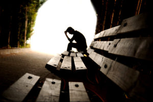 A person sits on a bench and appears to be upset as they lay their head into their hands.