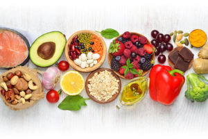 A wide variety of diet rich foods are shown; including, but not limited to, a red bell pepper, lemon, avocado, berries, nuts and garlic.