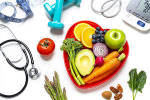 A red heart-shaped plate is filled with vegetables and fruit. The plate is surrounded by medical equipment.