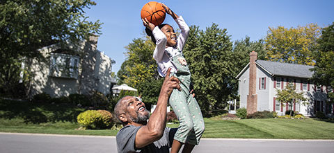 Aldo Williams lifts his granddaughter up as she holds a basketball over her head in a driveway.