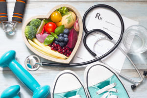 A stethoscope, a pair of blue dumbbell weights, a pair of blue tennis shoes and a heart-shaped bowl full of fruit is shown. 